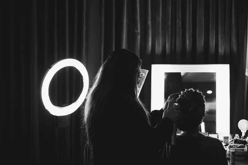 Black and white photo of a hairdresser styling a woman's hair in a salon with a ring light and vanity mirror