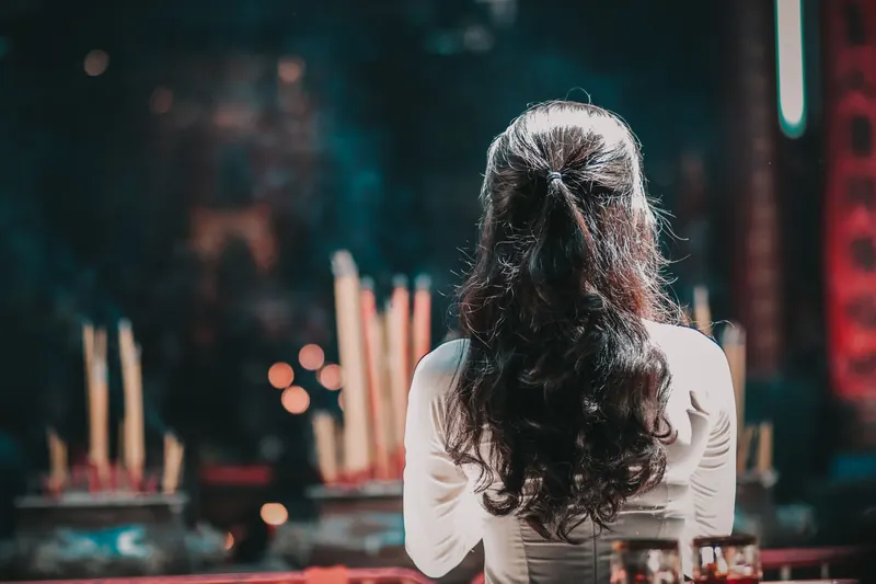 A woman with long hair is seen from behind praying inside a Vietnamese temple, surrounded by incense