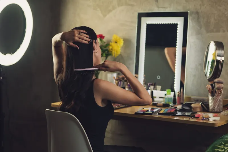 A woman styles her hair at a vanity with a mirror and various cosmetics, creating a personal grooming routine indoors