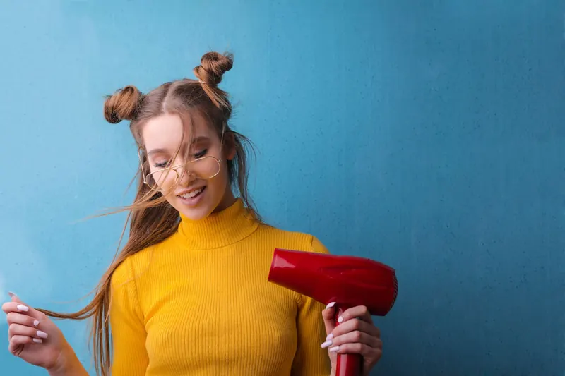 Woman with quirky hairstyle and glasses smiling with a hair dryer against a blue background