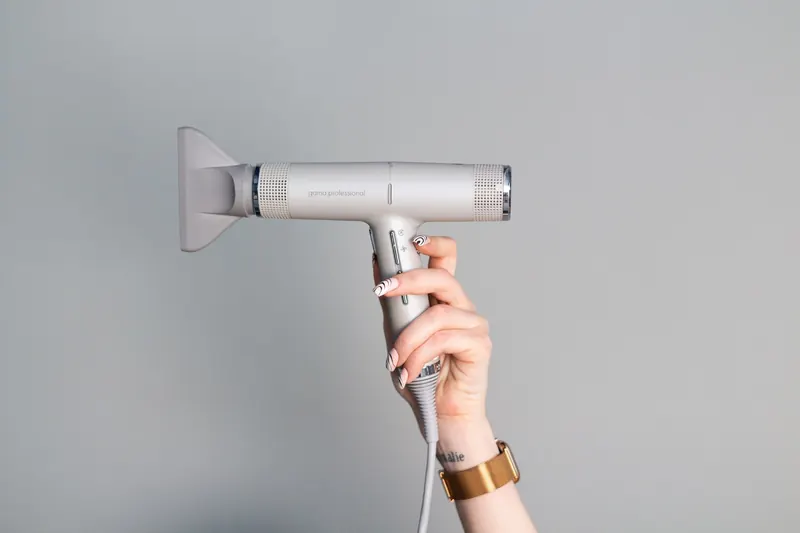A close-up of a hand with manicured nails holding a sleek hair dryer against a neutral background