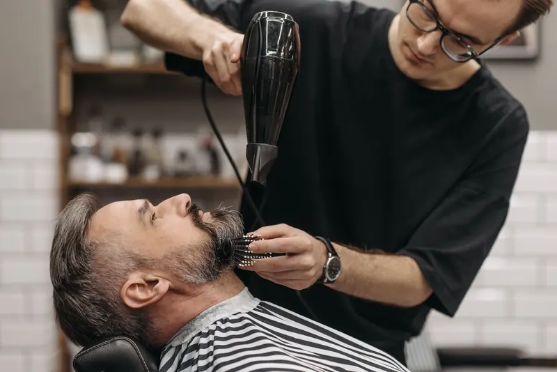 Barber styling a man's beard with hair dryer in a modern salon, capturing the art of grooming
