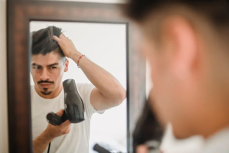 Adult man styles his hair using a hairdryer while looking at his reflection, capturing a modern grooming routine