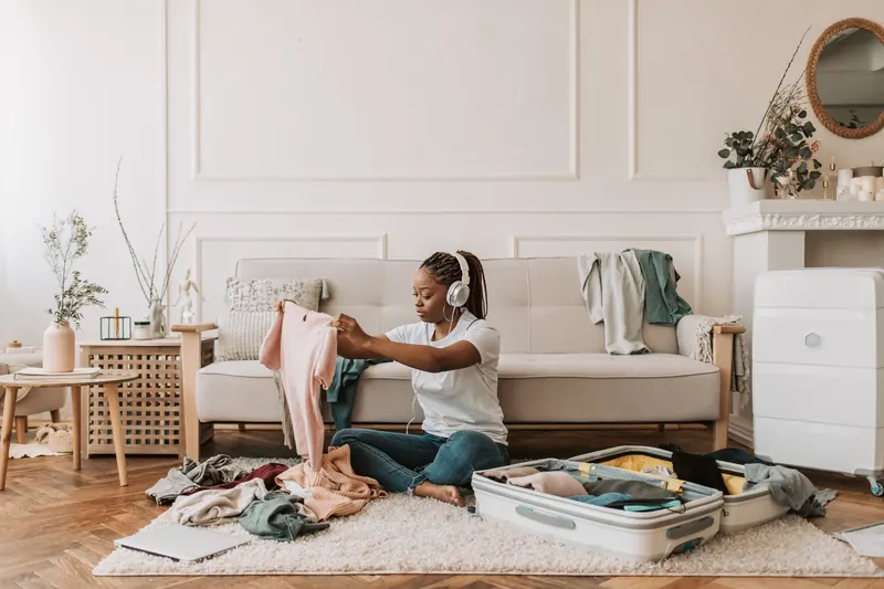 A woman with braided hair packs a suitcase at home while listening to music on headphones