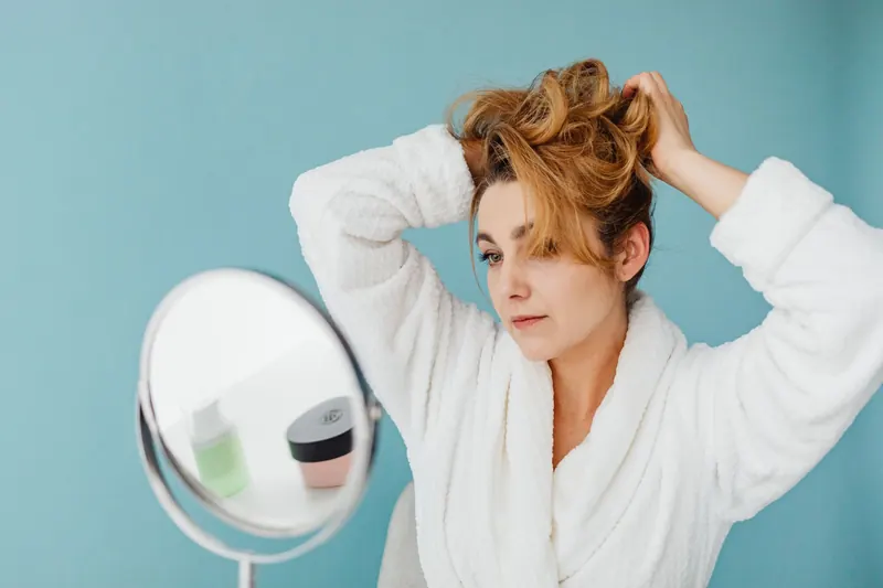Woman in bathrobe looks in mirror while styling hair, in a modern indoor setting