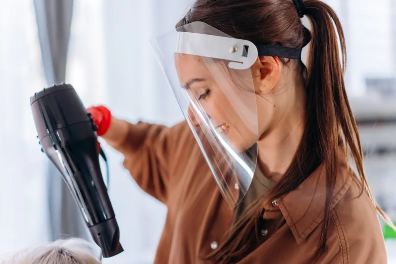Hairdresser wearing a face shield, using a hair dryer to style hair. Indoor salon setting with a safety focus
